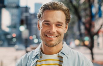 A man with light brown hair and a beard smiles at the camera, wearing a light denim jacket over a yellow and white striped shirt, with an urban street background.