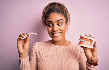 An Afro-Amarican young woman holiding a dental model with metal braces in one hand and an Invisalign aligner in the other