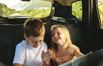 Two young children, a boy and a girl, are sitting in the backseat of a car, laughing together during a car ride.