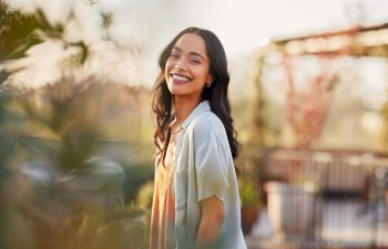 A woman with long dark hair smiles outdoors on a sunny day, standing amid greenery with a blurred background.